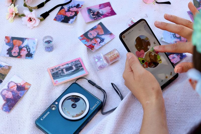 Hands holding a smartphone displaying the Canon Mini Print app, with mini prints scattered all around. 