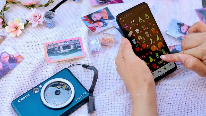 Teal Canon Zoemini S2 instant camera and printer surrounded by printed stickers in a tent with a person holding smartphone showing Mini Print App with different stamps.