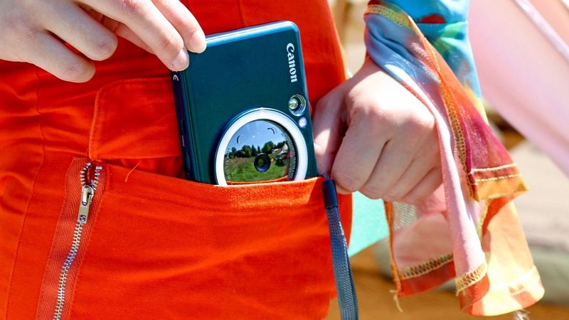 Teal Canon Zoemini S2 instant camera and printer being put in a pocket of brightly-coloured trousers.
