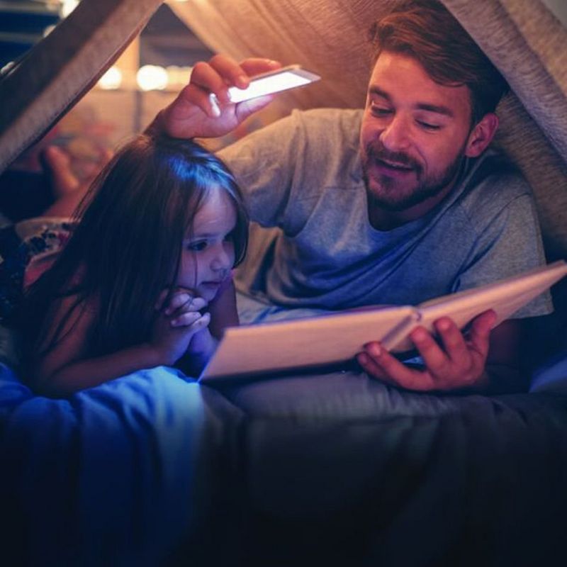 Man reading book to daughter in a tent at home using phone light