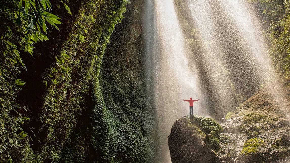 Man with his arms outstretched stood on a rock underneath a waterfall 