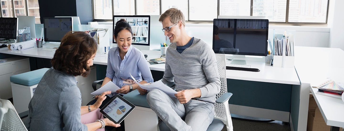 Two women and a man sitting in front of a long white desk adorned with PCs compare notes between pieces of paper and a tablet device.