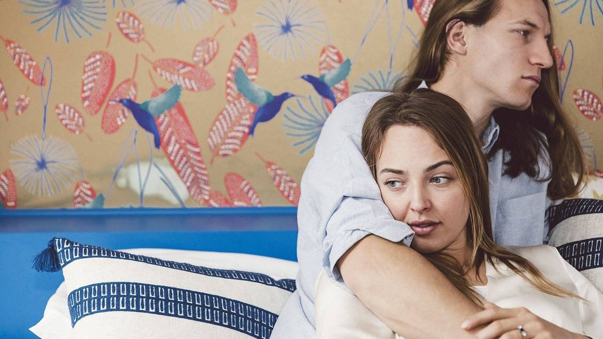 Male and female hugging on a bed in front of a floral wallpaper bedroom