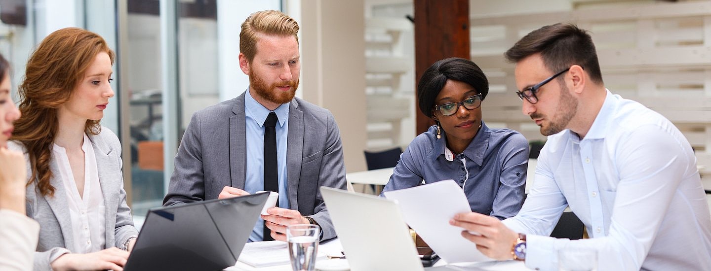 A group of men and women dressed in smart business attire sit round open laptops and a glass of water.