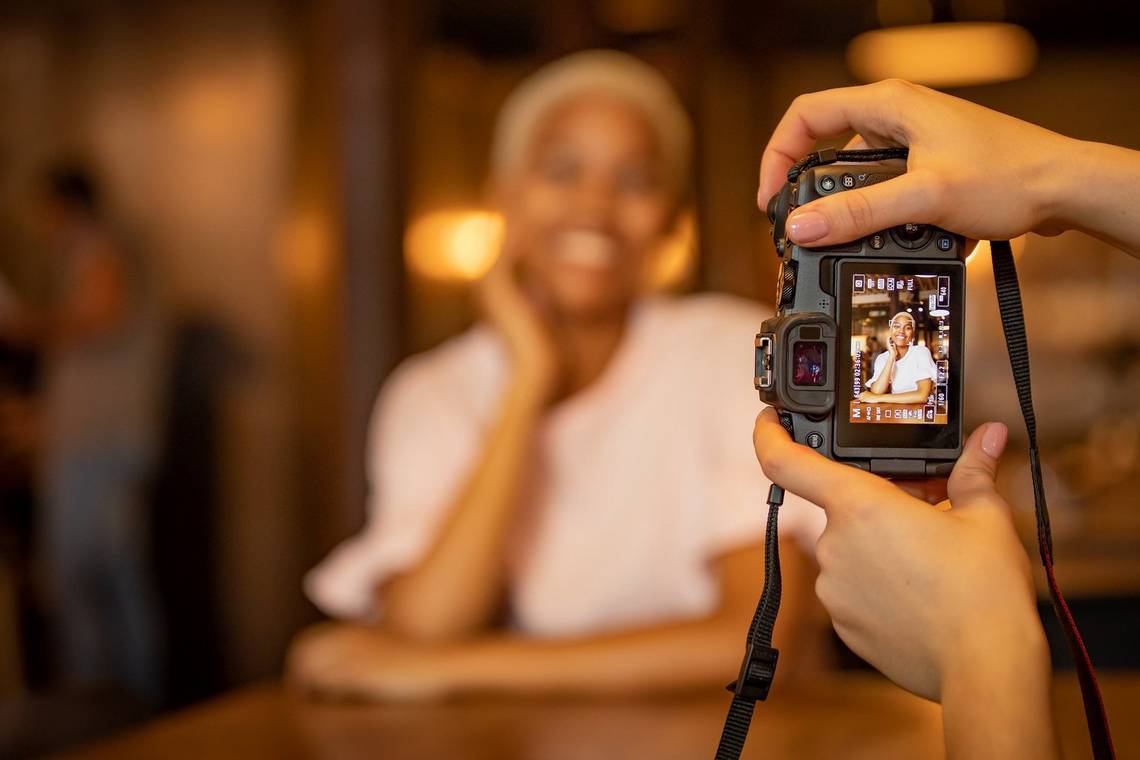  A photographer's hands hold a Canon йP as she photographs a smiling woman in a dimly-lit interior.