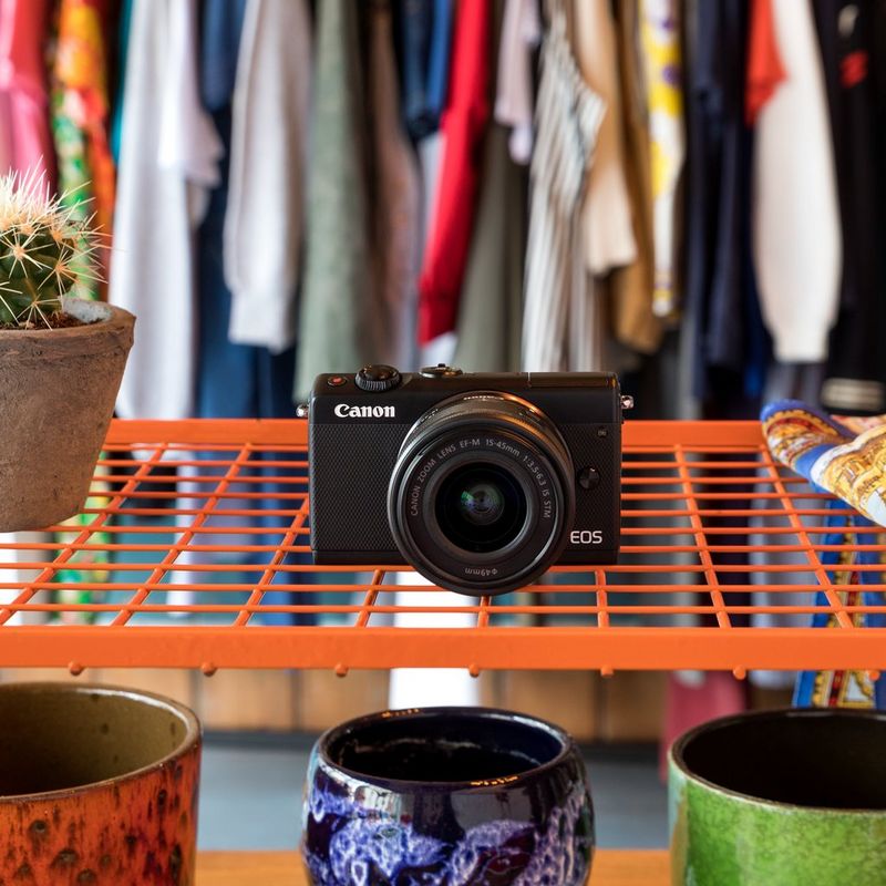 Canon EOS M100 Black + EF-M 15-45mm f/3.5-6.3 IS STM Lens Black Product Diagonal Back View with Built-in Flash and Tilted Screen