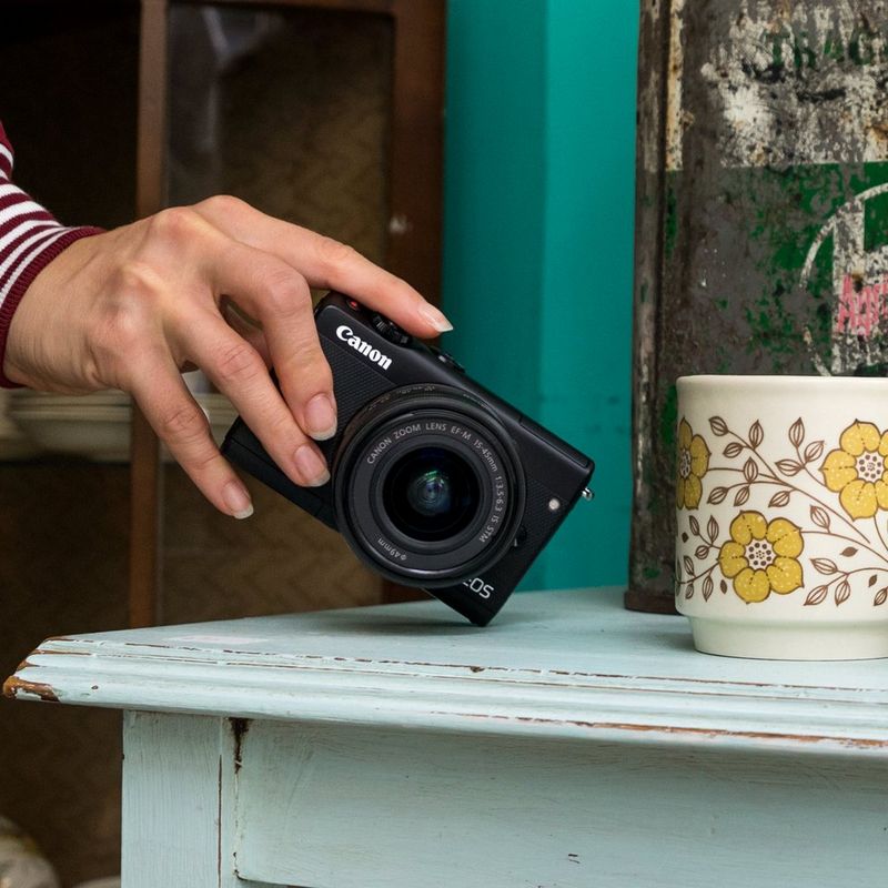Canon EOS M100 Body - Black  En hånd som plukker opp kameraet