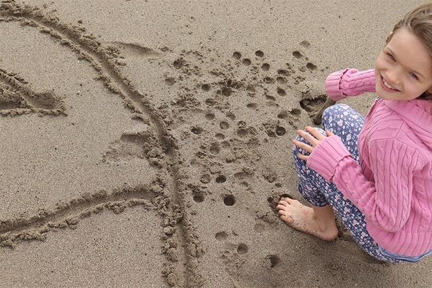 A smiling young girl on a beach, next to patterns she has drawn in the sand.