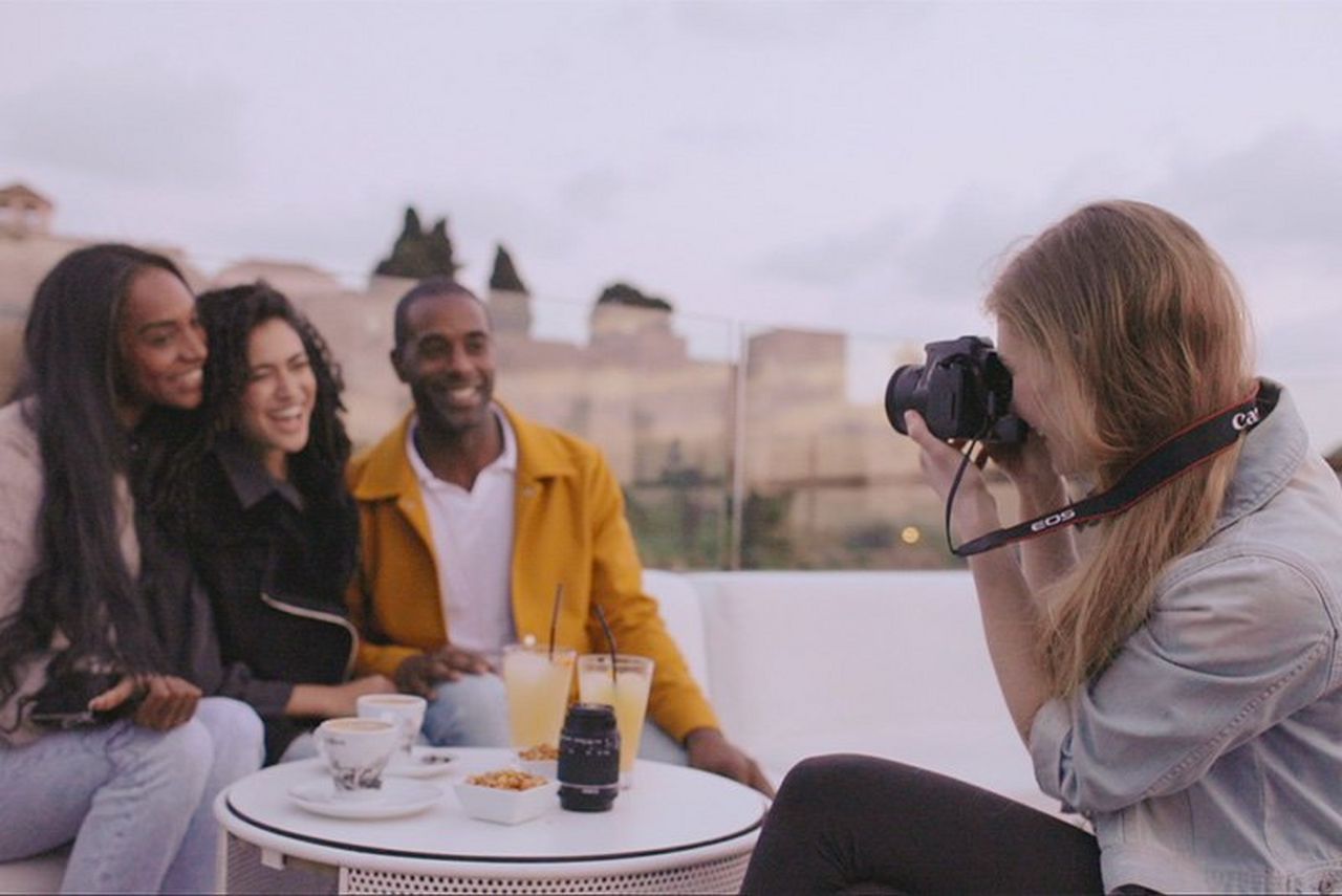 A woman with a Canon EOS 250D takes a portrait of three smiling people at a table on a patio outdoors.