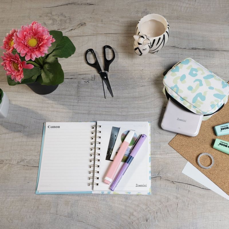 Impressora Fotográfica a Cores Portátil Canon Zoemini, Branco + Diário + Bolsa Impressora posicionada numa secretária ao lado de umas tesouras