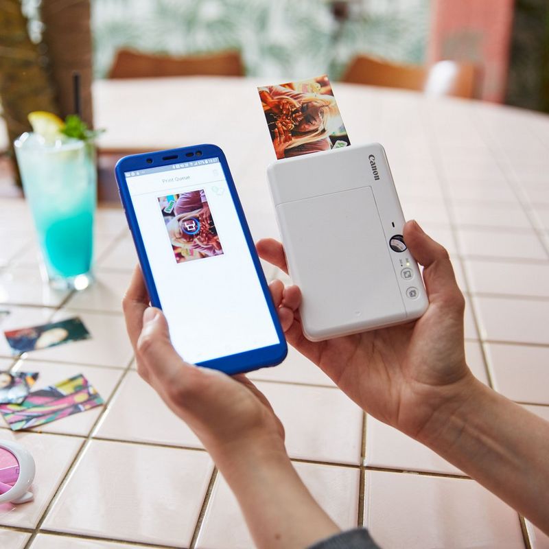 Cámara impresora instantánea Canon Zoemini S, blanco perla Impresora con una impresión saliendo y una mano con una cámara