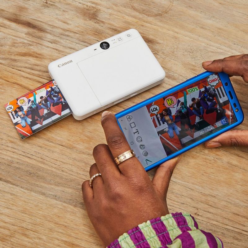 Cámara impresora instantánea Canon Zoemini S, blanco perla Una cámara y una impresora con una impresión saliendo sobre un escritorio