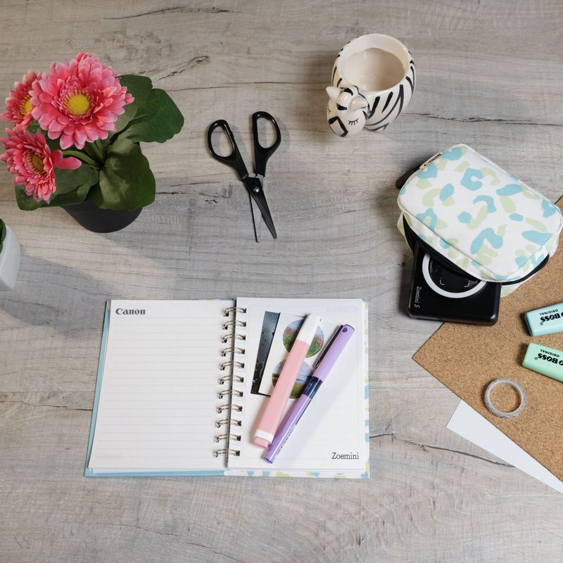Cámara impresora fotográfica en color instantánea Canon Zoemini S, negro mate + Diario + Estuche Impresora colocada sobre un escritorio con tijeras