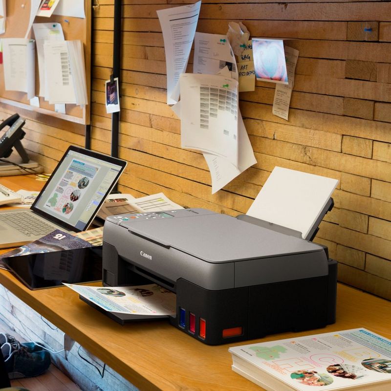 Canon PIXMA G3520: impresora MegaTank 3 en 1 inalámbrica en color con depósitos de tinta rellenable Sobre una mesa de oficina junto a un portátil con la bandeja de papel abierta y un soporte de papel