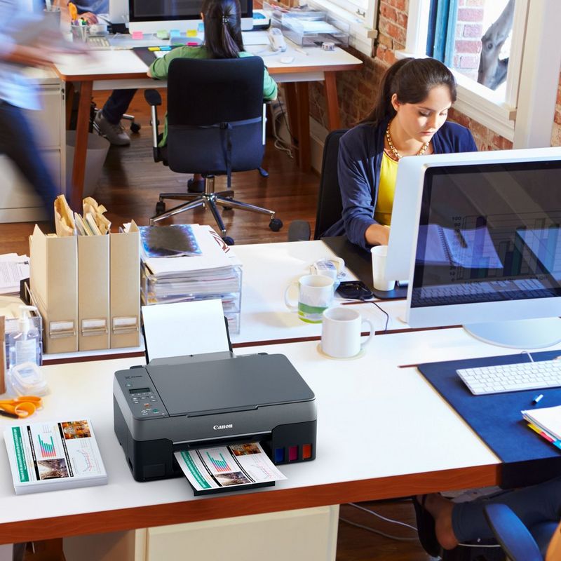Canon PIXMA G3560: impresora MegaTank 3 en 1 inalámbrica en color con depósitos de tinta rellenable Sobre una mesa de oficina junto a un escritorio con la bandeja de papel abierta y un soporte de papel