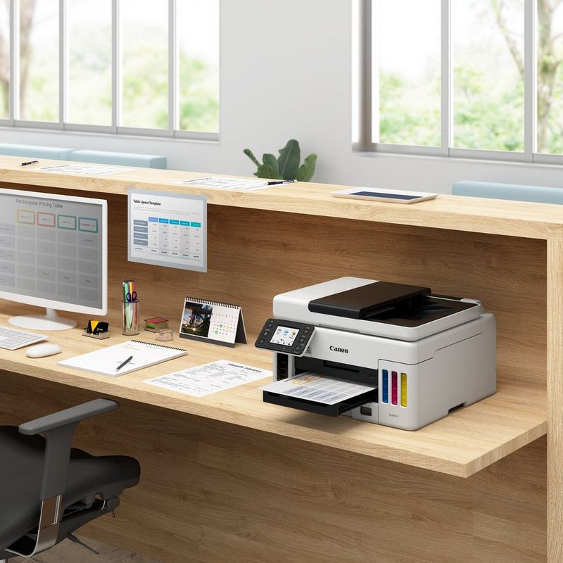 Canon MAXIFY GX6050 Wireless 3-in-1 Refillable MegaTank Inkjet Printer  På et kontorbord ved siden av en stasjonær datamaskin med papirskuff åpen