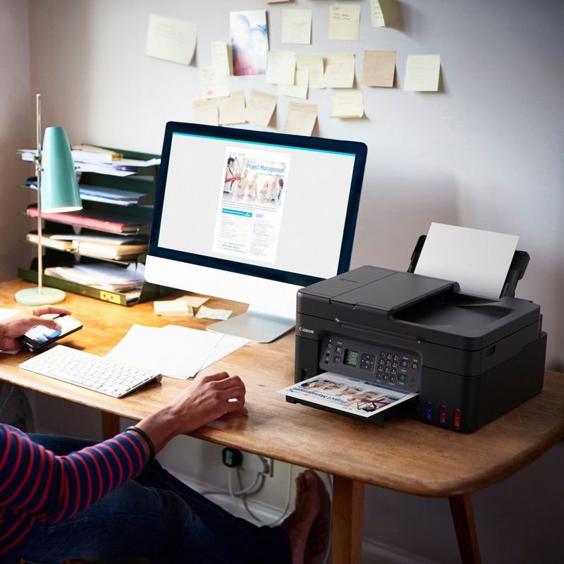 Canon PIXMA G4570: impresora multifunción inalámbrica MegaTank de inyección de tinta color con depósitos de tinta rellenables Producto en el escritorio de una oficina junto a una mujer sujetando un teléfono