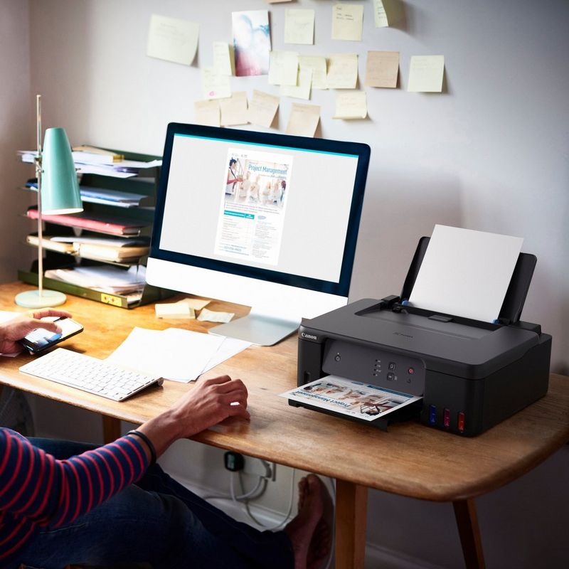 Imprimante MegaTank rechargeable couleur Canon PIXMA G1530 Produit posé sur un bureau avec une femme tenant un téléphone