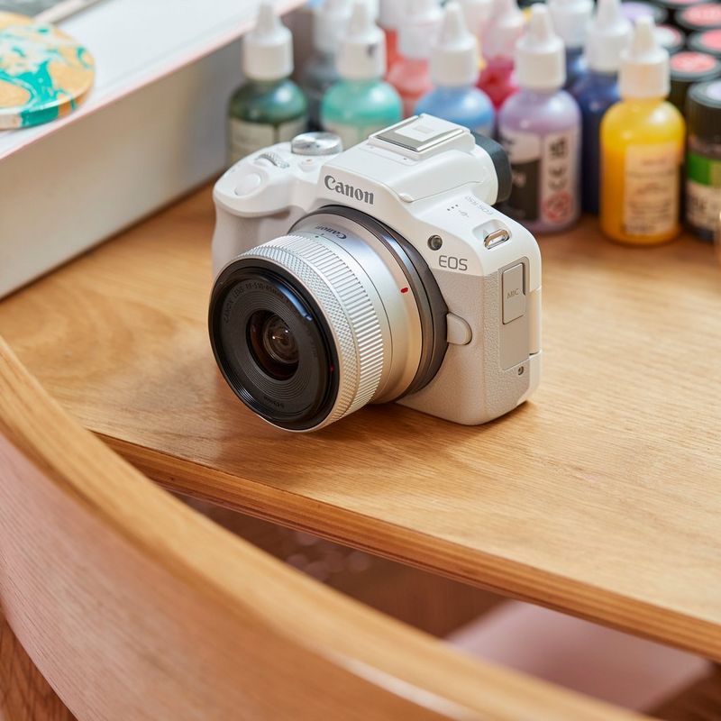 Canon EOS R50 Mirrorless Camera, White and RF-S 18-45mm F4.5-6.3 IS STM Lens Product on a Desk with Color Bottles