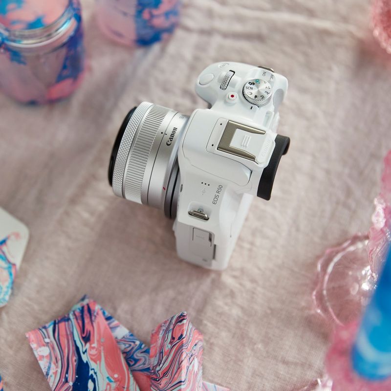 Canon EOS R50 Mirrorless Camera, White and RF-S 18-45mm F4.5-6.3 IS STM Lens Product kept on a table with Pink Cloth
