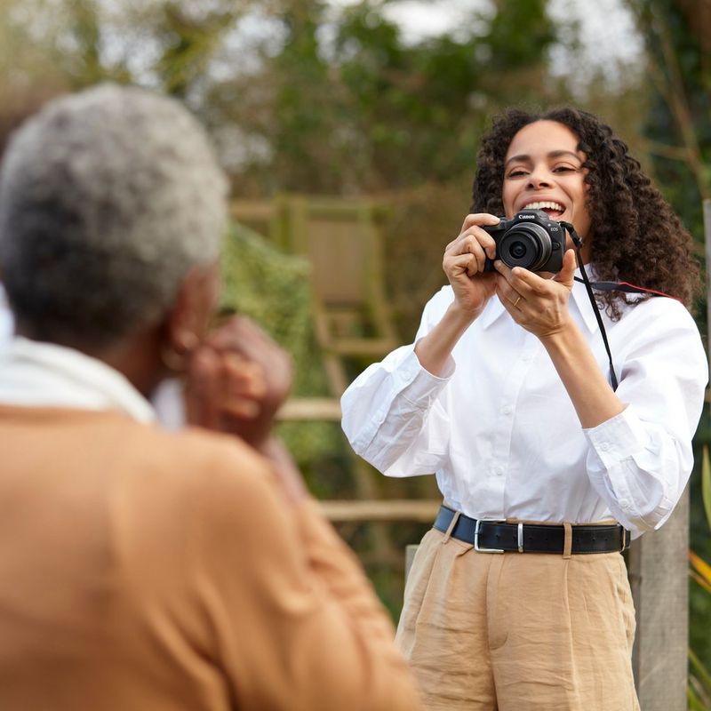 Canon EOS R100 Mirrorless Camera Woman holding product with neck strap taking photo of man