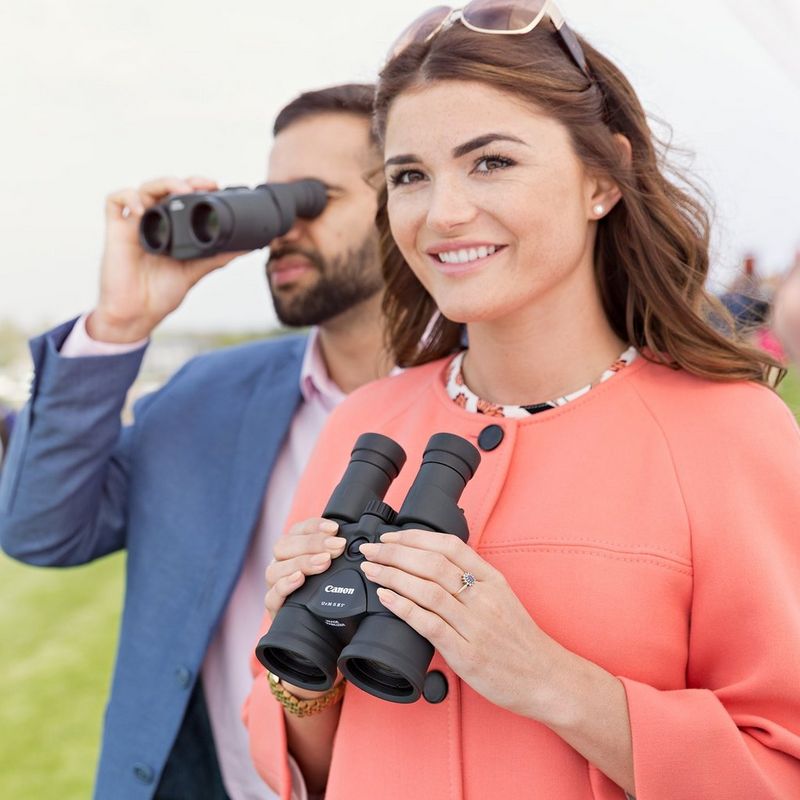 Canon 12x36 IS III Binoculars Man in the Background Using Binoculars and Woman in the Foreground Smiling and Looking Ahead Holding the Binoculars Near her Face
