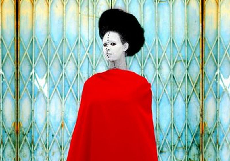 A woman stands in front of a rusting security grille. She wears a dramatic red cape, which covers her from the neck down, and her head is framed by a halo of black hair. Her face is painted white and a line of black dots stretches from the top of her forehead to the base of her neck, which are inspired by traditional African body art.