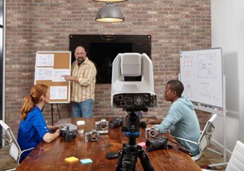 A man with a beard stands by a pin board, pointing at what is pinned on it. He wears a yellow checked shirt and blue jeans. In front of him, with their backs slightly turned to the camera, are two people – one on each side of a brown wooden table, upon which are assorted camera bodies and a coffee cup. On the left is a woman with red hair and a bright blue blouse. On the right is a man with a pale blue hoodie. In the centre of the shot, at the head of the table, is the rear of a Canon PTZ camera.