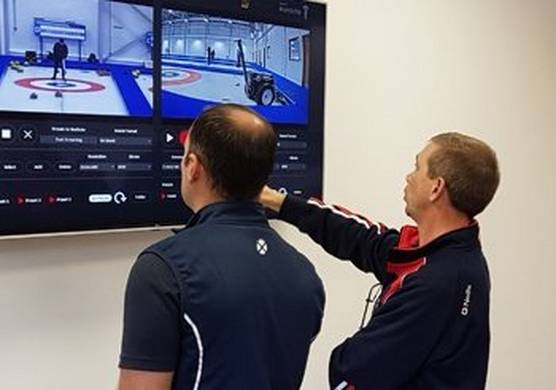 Two men in athletic wear look at a split screen showing two views of curling players on the ice.