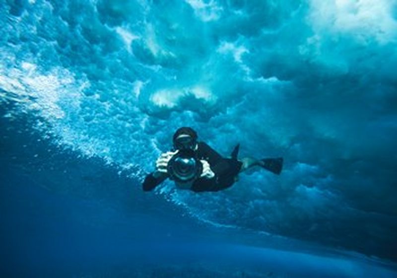 A person swimming under the sea, wearing goggles, a wetsuit and flippers. They are holding an underwater camera and pointing it directly at the camera. Behind them is the seafoam above.