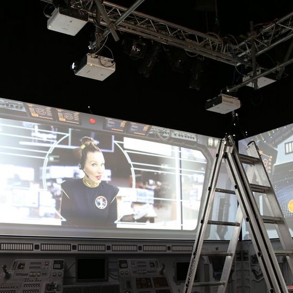 A ladder and projectors pointing to a screen, where a woman in a headset is broadcast.