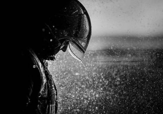 A black and white photo of a man in a helmet. He stands to the left of the image, with rain pouring down in front of and on him.