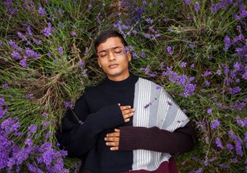 A bespectacled young man in a black and grey sweater lies in long grass, surrounded by bluebells. His right-hand rests on his chest at heart height. (© Boys by Girls Magazine)
