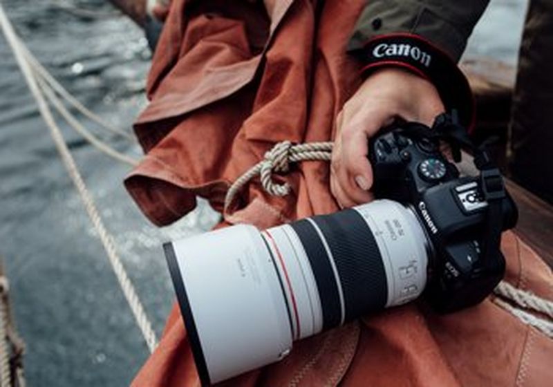 A Canon camera with a single hand holding it as it rests on an orange tarpaulin. Besides the tarpaulin are ropes and beyond them is the sea. Together it appears that the photographer is on a boat at sea.