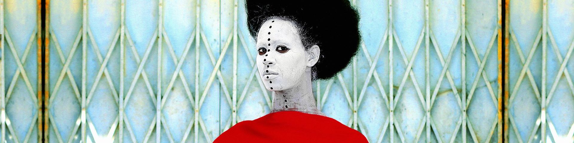 A woman stands in front of a rusting security grille. She wears a dramatic red cape and her head is framed by a halo of black hair. Her face is painted white and a line of black dots stretches from the top of her forehead to the base of her neck, which are inspired by traditional African body art.