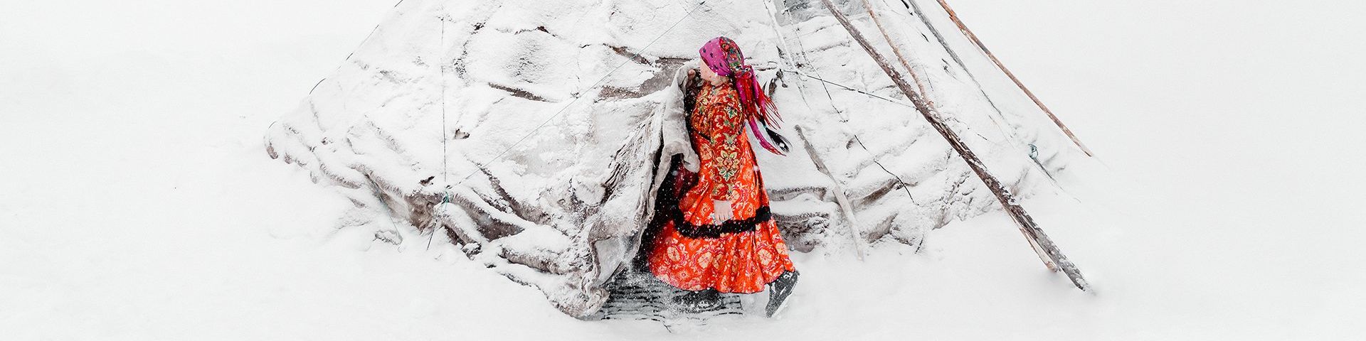 The image is dominated by pure white snow, save a snow-covered chum, or traditional tent used by the migrational tribes of the region. A woman emerges from her tent and is wearing a long orange/red patterned dress and boots. The dress has a black trim midway up the skirt and she also wears a pink headscarf, tied at the back of her head, which swings behind her.