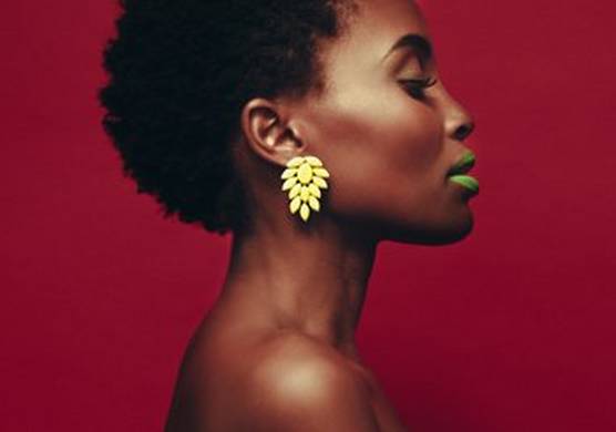 The head and neck of a dark-skinned woman is photographed from the side against a deep burgundy background. Her hair is short, exposing her ear and a large yellow floral-style drop earring. Her lips are painted green.