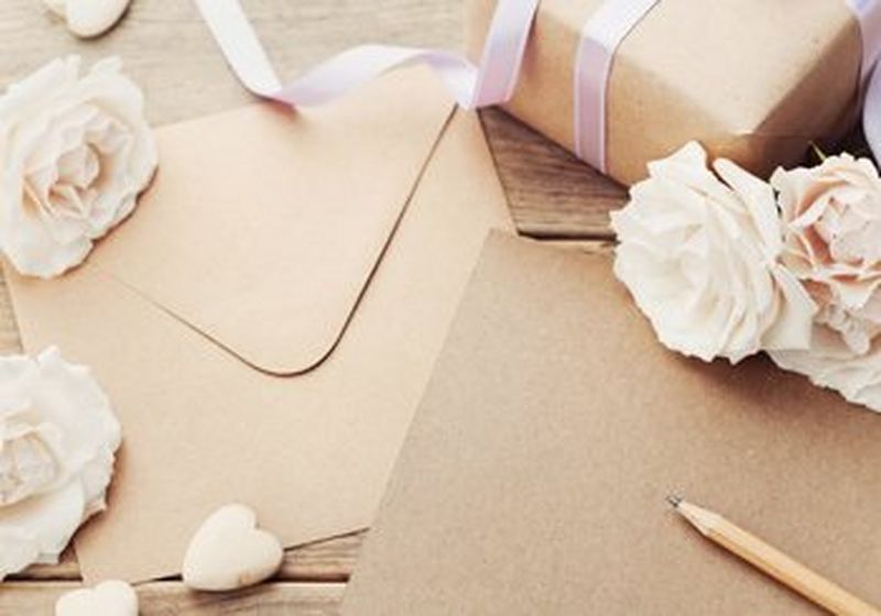 A brown paper envelope and the edge of a brown kraft paper card, on a table, surrounded by white rose-like flowers.