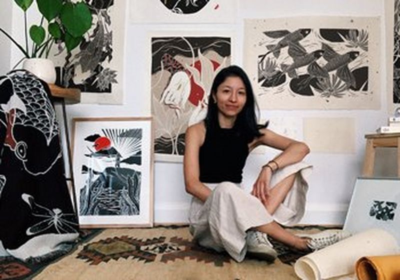 A woman sits on the floor surrounded by fine art prints in the Japanese style. They predominantly in black, grey and red. She has shoulder-length dark hair and wears a beige skirt, white sneakers and a bleck vest-style top. On the left and right of the shot are two tables, one with a green plant on top and a black and white printed textile in front. The other with some books stacked on top and prints in front of it.