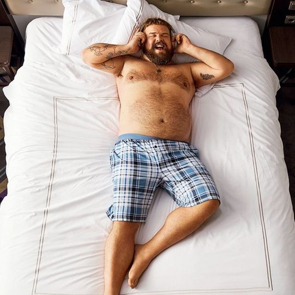 A bearded man lies on a white bed wearing blue checked shorts. He is talking on the phone and laughing.