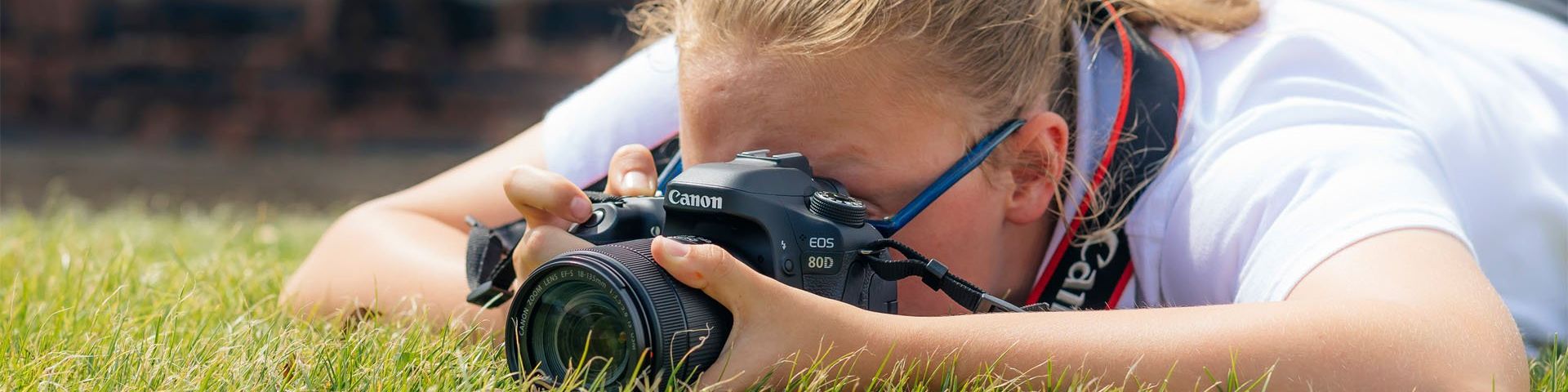 A girl lies on the grass, her face obscured by a Canon EOS 80D, as she takes a shot