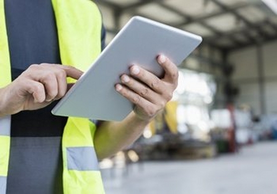 The body of a man in a high-vis jacket, he stands in a warehouse holding a tablet computer.