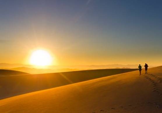 On the right, a small silhouette of a couple walking across golden sand dunes at dusk, leaving footprints behind them. Far on the horizon, a huge bright sun begins to set.