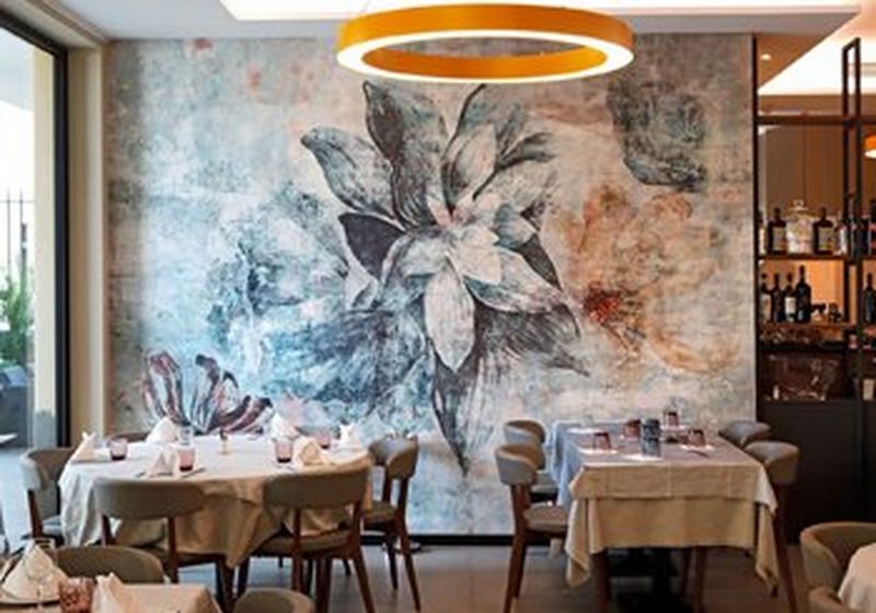 A restaurant setting, with two tables side by side. One is round, with six chairs. The other is oblong with four chairs. Both are set ready for customers, with cream tablecloths. Above them is a circular orange light fitting and behind is an image of a huge flower, taking up the entire wall on printed wallpaper.