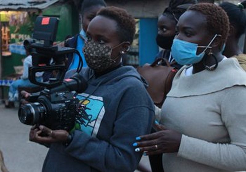 Two women – Jane Wanjiru Mumbi and Mercy Owegi. Both are wearing face coverings and only with head and shoulders visible, are facing left as though looking at something. The woman on the furthest left holds a Canon video camera. Behind them stand two further women.
