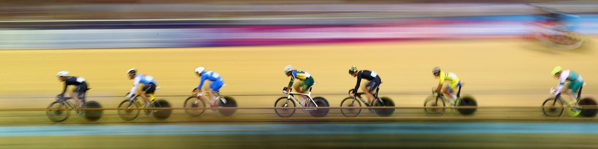 A photograph of seven professional cyclists in a line along what must be a velodrome. However, the photograph has substantial motion blur and all that can be seen of the background are streaks of yellow, blue and pink.
