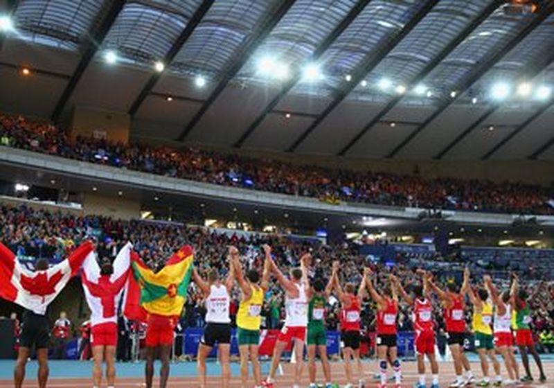 Athletes line up on the track to face the stadium crowd. They hold hands and raise them above their heads. Some are wearing their nations flags on their shoulders. Others simply bear their bib numbers. 