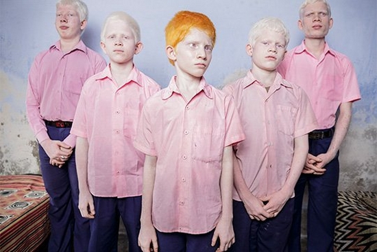 Five boys in pink shirts stand in a V-shaped formation in a blue room. Four of them have very pale skin with blonde hair, one has pale skin with red hair. All have pale eyes.