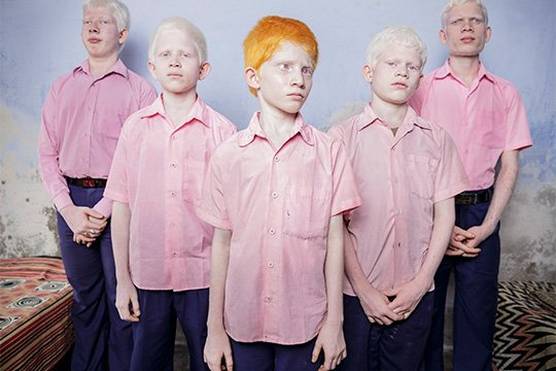 Five boys in pink shirts stand in a V-shaped formation in a blue room. Four of them have very pale skin with blonde hair, one has pale skin with red hair. All have pale eyes.