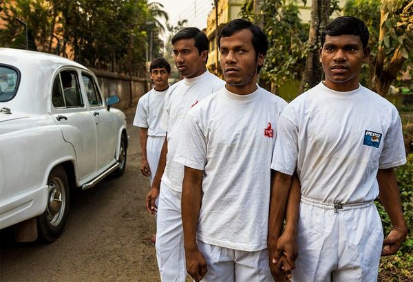 Four young men in white t-shirts and white trousers walk beside a road holding hands as a car passes. Some are visibly blind.